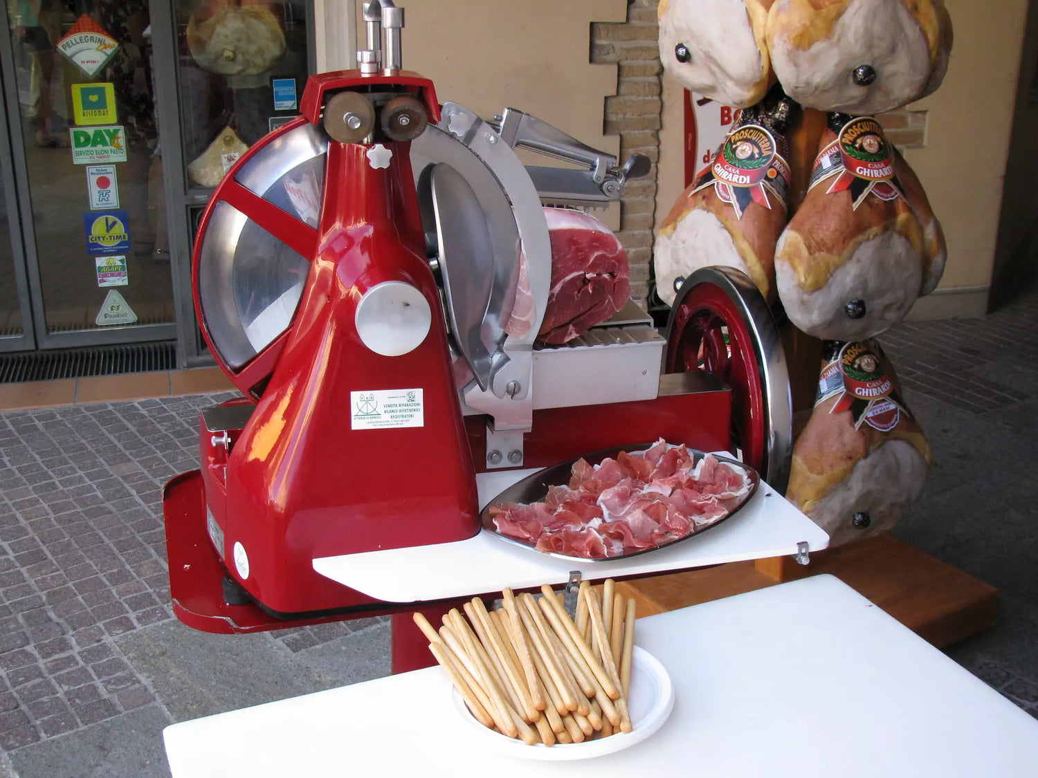 Affettatrice rossa in azione: prosciutto tagliato sottile, accanto a croccanti grissini su un tavolo in un'ambientazione rustica.