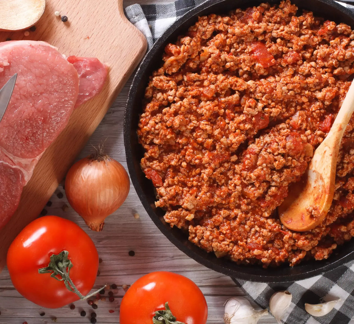 Deliciosa salsa de carne piamontesa hirviendo a fuego lento en una sartén negra, acompañada de tomates, cebollas y finas lonchas de carne dispuestas sobre una tabla de cortar de madera.