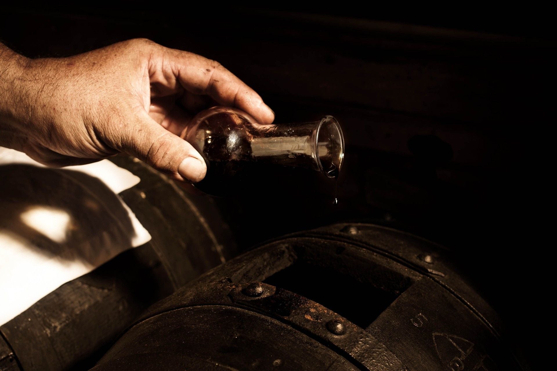 Una mano sta versando Aceto Balsamico Tradizionale di Modena DOP proveniente da una minuscola anfora su barili di legno antichi.