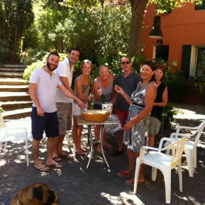 Gruppo di persone allegre all'aperto con una torta su un tavolo, in giardino circondato da piante verdi.