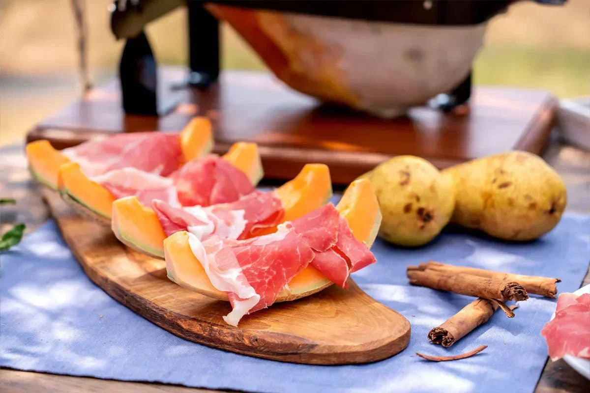 Prosciutto di Parma su melone e tavola rustica, con pere e cannella, evoca sapori estivi italiani autentici e freschi.
