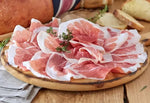Tagliere di legno che presenta fette rosate di Prosciutto di Parma DOP, stagionato 18 mesi, disossato e arricchito con erbe fresche.