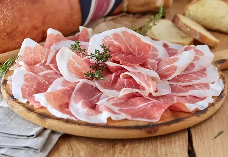 Tagliere di legno che presenta fette rosate di Prosciutto di Parma DOP, stagionato 18 mesi, disossato e arricchito con erbe fresche.