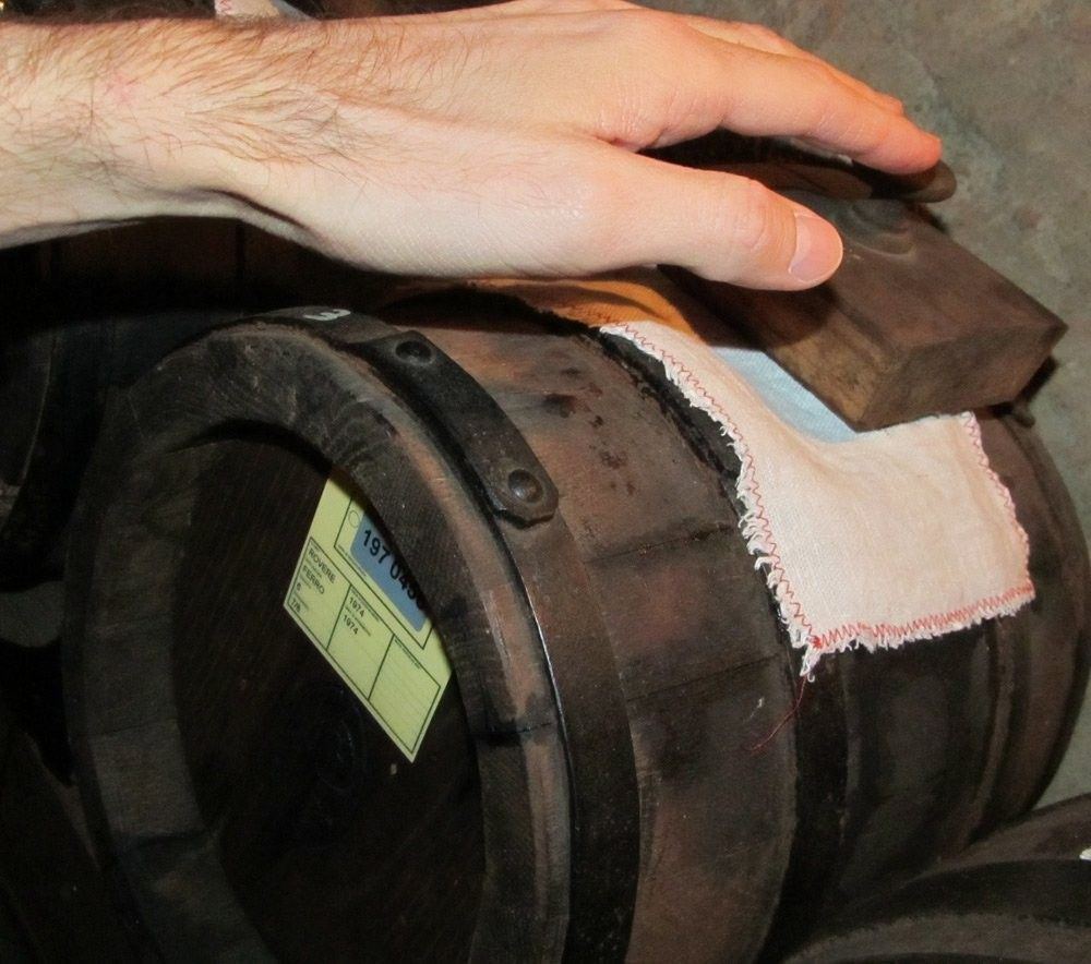 Atmosfera rustica pervade mentre una mano poggia su una botte antica di legno, coperta da stoffa bianca, simbolo di tradizione e qualità artigianale per l'aceto.