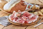 Deliziose fette di Prosciutto di Parma DOP, stagionato per 18 mesi, morbide e dal colore rosato, presentate su un tagliere. In secondo piano, si scorge il prosciutto intero con osso.