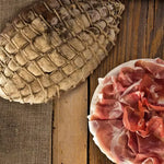Un intero Culatello Tradizionale, avvolto in rete, è posato su una tavola rustica di legno, con accanto affettati rosati. Ha una stagionatura di oltre 12 mesi.