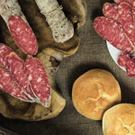 Un tagliere di legno presenta un rustico salame di Felino IGP affettato, servito con croccante pane, evocando sapori italiani autentici.