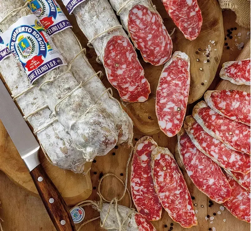 Un rustico salame di Felino IGP si adagia su una tavola di legno, circondato da sfumature rosse e bianche, con un coltello affilato a fianco.