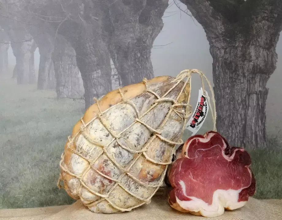 Un intero Fiocchetto di Prosciutto, stagionato per 6 mesi e appeso con una corda, si presenta succulento su uno sfondo nebbioso di alberi.