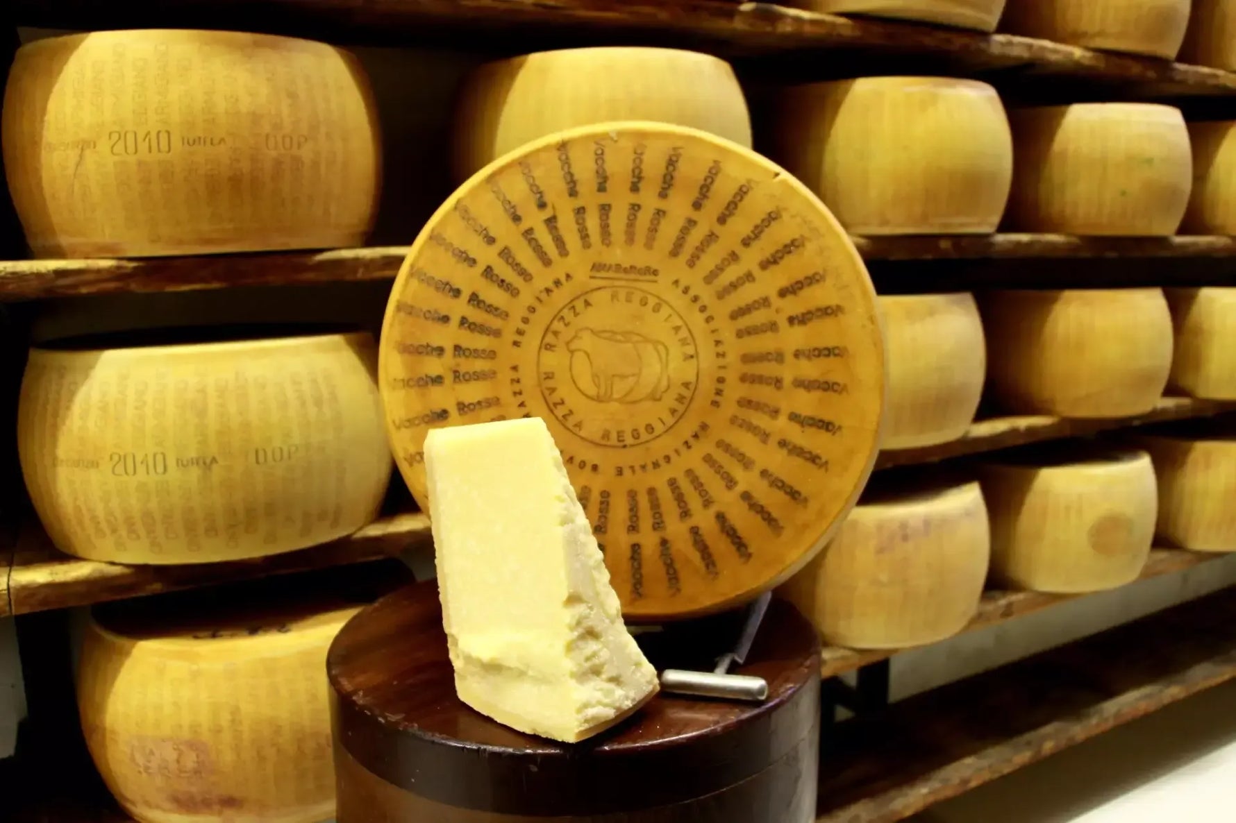Un'intera forma di Parmigiano Reggiano Vacche Rosse, con 24 mesi di stagionatura, presenta colori dorati maturati e riposa su scaffali in legno.