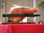Una morsa per prosciutto, realizzata in acciaio lucido, posa su una base di legno, unendo eleganza e funzionalità per un'affettatura precisa.