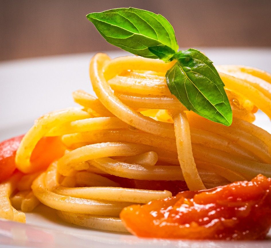 Squisiti spaghetti di grano duro, delicatamente avvolti in una salsa di pomodoro, arricchiti da una fresca foglia di basilico, serviti su un piatto bianco.