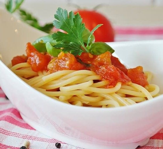 Un piatto bianco di spaghetti di semola, conditi con salsa di pomodoro fresco, basilico e prezzemolo, presentato su una tovaglia a righe.