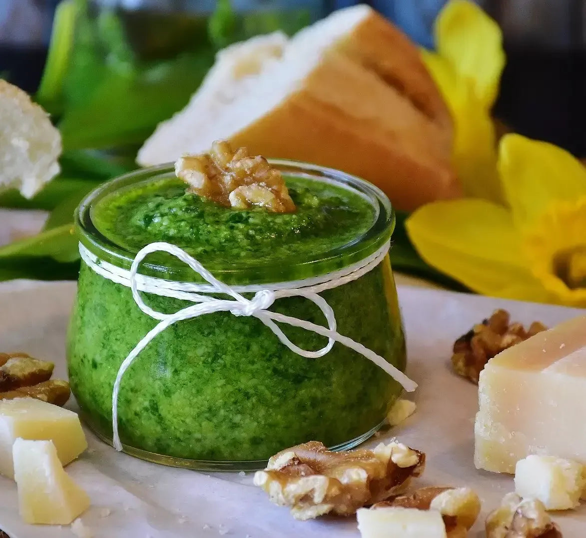 Un barattolo di pesto al basilico, legato con un nastro, è incorniciato da noci, formaggio e pane, su un pittoresco sfondo di foglie verdi e narcisi fioriti.