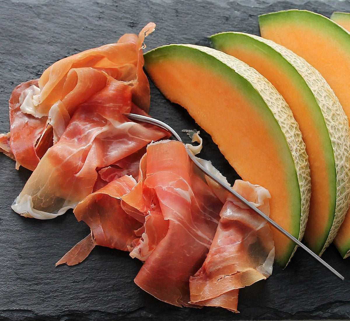 Vue d'un jambon de Parme AOP, affiné pendant 24 mois, accompagné de tranches de melon sur une assiette en ardoise foncée.