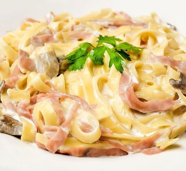 En hvit tallerken med økologiske eggpappardelle, tilberedt med en krem av sopp og skinke, og beriket med fersk persille.