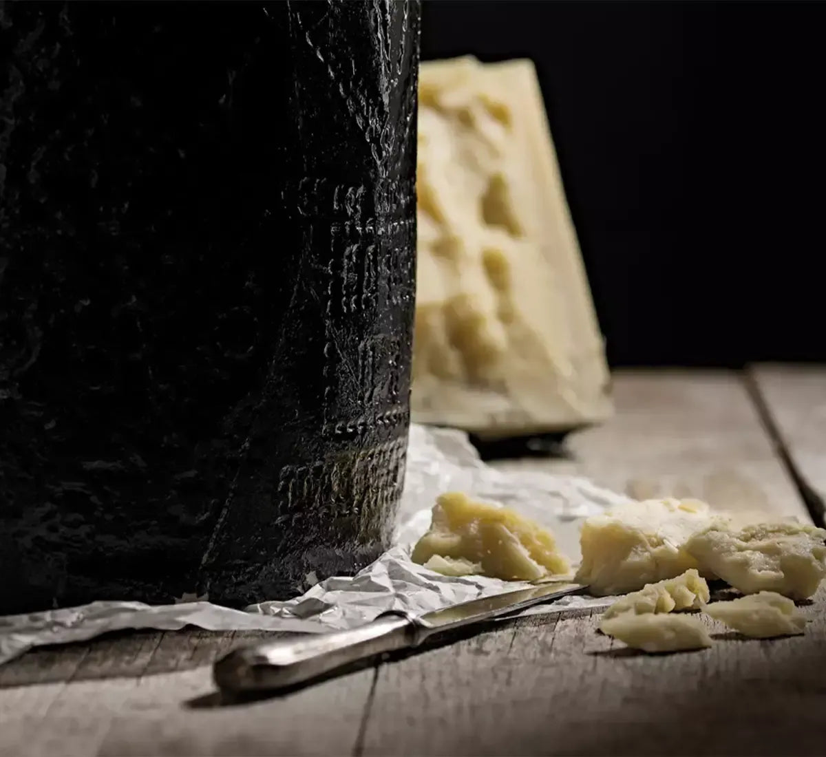 Sullo sfondo rustico di una tavola, un Pecorino Romano DOP stagionato, con un coltello e scaglie aromatiche vicine.