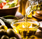 Un olio d'oliva dorato, della marca Signore degli Ulivi, viene versato in un recipiente di vetro, affiancato da olive fresche e insalata.