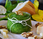 Un vasetto di pesto verde fatto a mano, arricchito di pinoli e formaggio, è affiancato da pane fresco, con un tocco di fiori gialli sullo sfondo.