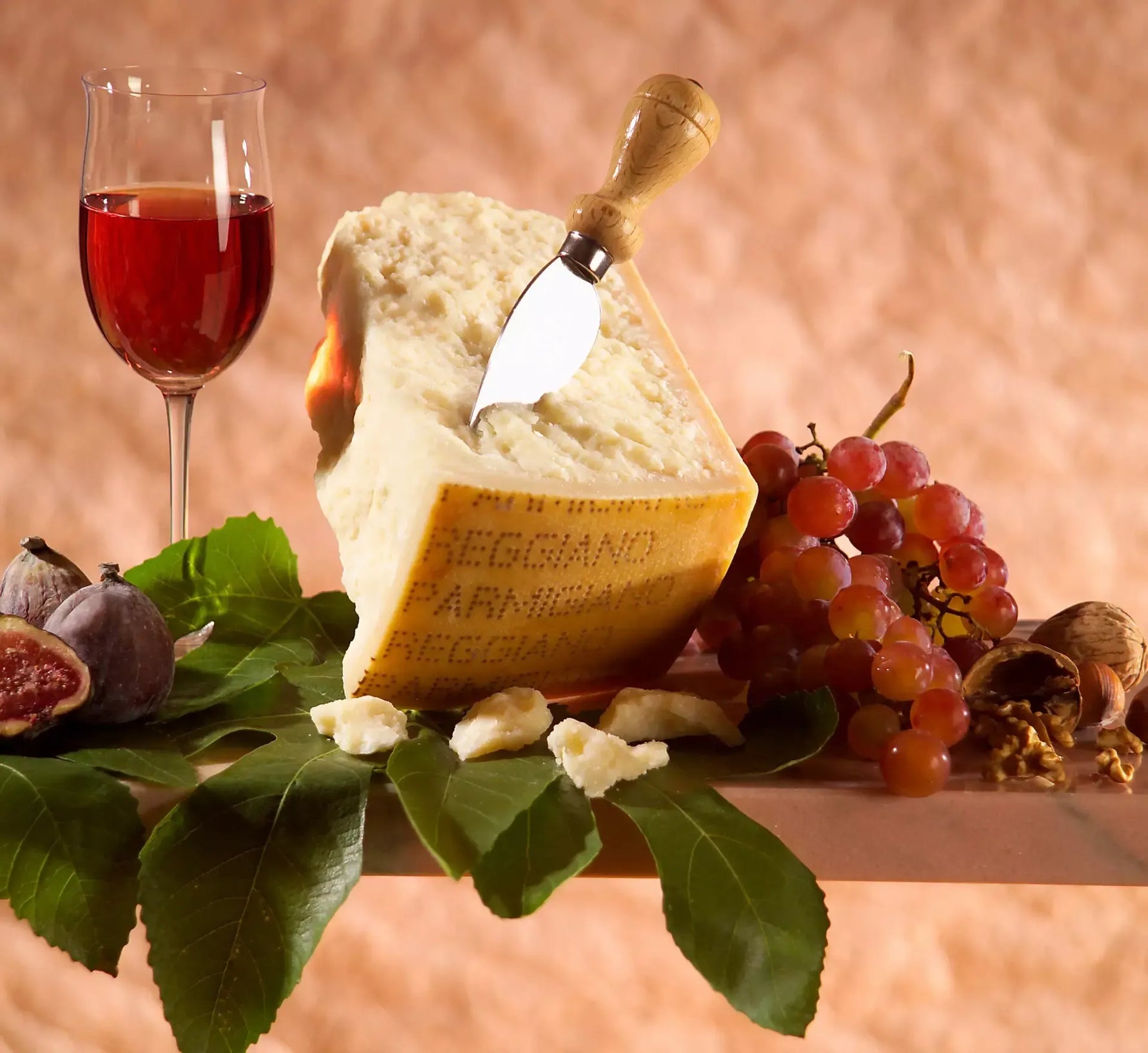 Un pregiato Parmigiano Reggiano di montagna stagionato 36 mesi, accompagnato da coltello, grappe, fichi succulenti e un bicchiere di vino rosso, il tutto presentato su un letto di foglie verdi.