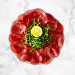Un piatto elegante in marmo bianco ospita fette rosse di bresaola, arricchite da rucola e un tocco di limone.