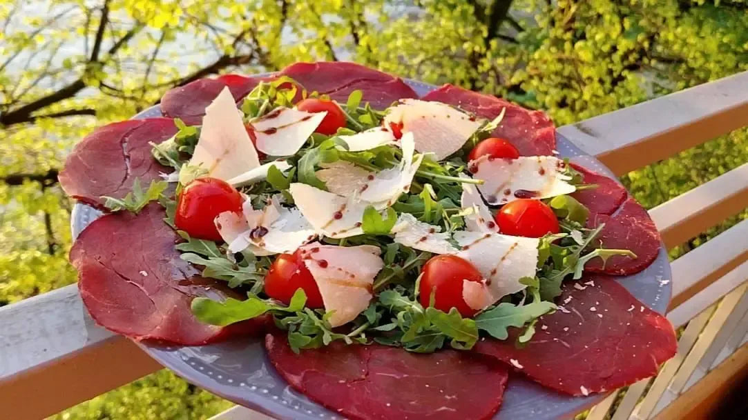 De délicieuses tranches de bresaola de bœuf, agrémentées de roquette, de tomates cerises et de copeaux de parmesan, présentées sur une table extérieure.