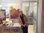 Un'affascinante donna, con un sorriso radioso, si trova in una formaggeria, presentando orgogliosamente il Parmigiano Reggiano Vacche Rosse, posizionata vicino a un poster vivace di una mucca rossa.