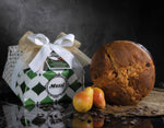 Un Panettone Pere & Cioccolato, delicatamente incartato a mano, è adornato da un nastro elegante. Accanto, pere fresche e scaglie di cioccolato lo completano.