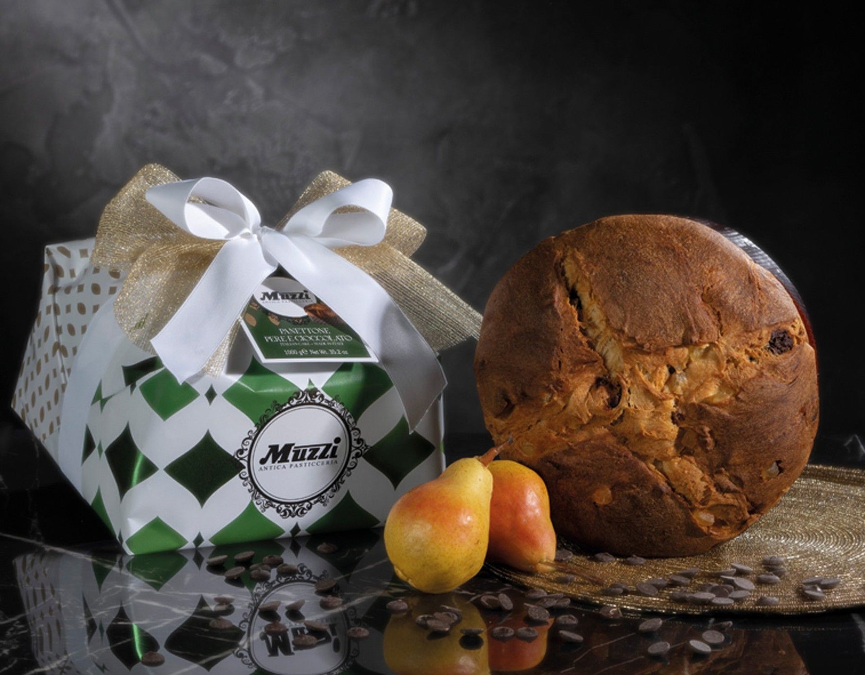 Un Panettone Pere & Cioccolato, delicatamente incartato a mano, è adornato da un nastro elegante. Accanto, pere fresche e scaglie di cioccolato lo completano.