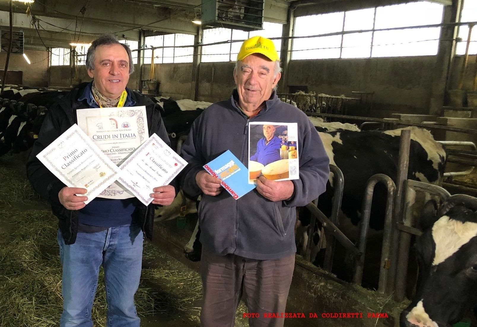 I produttori Bonati esibiscono con orgoglio certificati e un libro, circondati da mucche all'interno di una stalla.