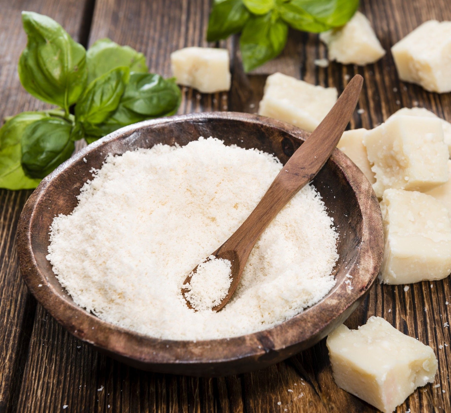 Una ciotola di legno colma di Parmigiano Reggiano grattugiato, accanto a pezzetti di formaggio e foglie di basilico, posa su un tavolo rustico.