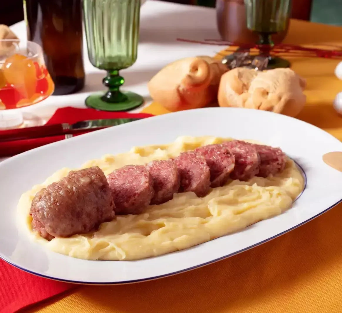 Un succulento cotechino precotto riposa su un cremoso purè di patate, accompagnato da bicchieri raffinati e panini croccanti: un elegante richiamo festivo.