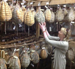 Una donna sta accuratamente scegliendo un Culatello di Zibello DOP stagionato, appeso come da tradizione, in una cantina, simbolo dell'eccellenza culinaria italiana.