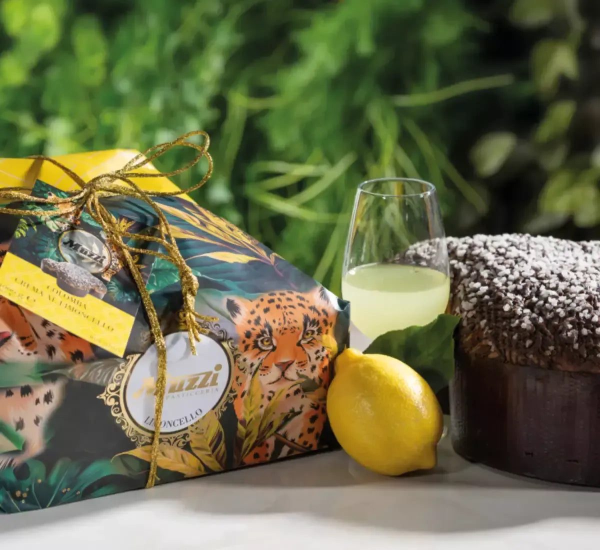 Una confezione di Colomba Limoncello, arricchita da un limone fresco, è accanto a un bicchiere di limoncello, il tutto immerso tra foglie verdi lussureggianti.