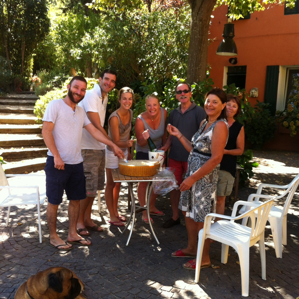 Gruppo di persone allegre all'aperto con una torta su un tavolo, in giardino circondato da piante verdi.