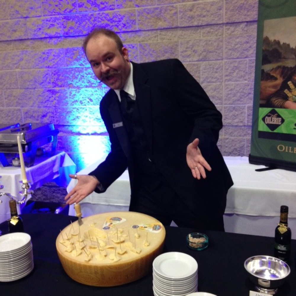 Un uomo elegante presenta un tavolo da formaggi gourmet in un evento raffinato, con bicchieri di vino e piatti pronti.